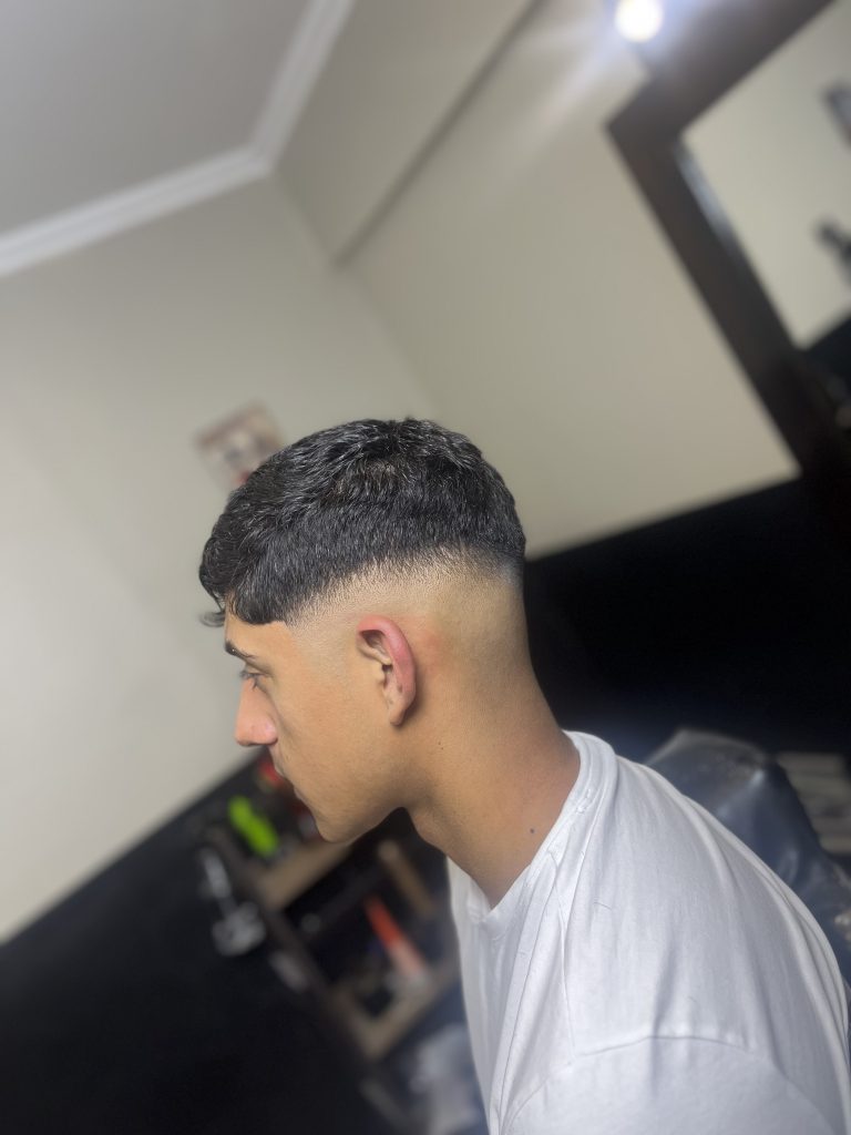Side profile of a man with a fresh skin fade haircut in a barbershop, wearing a white T‑shirt.
